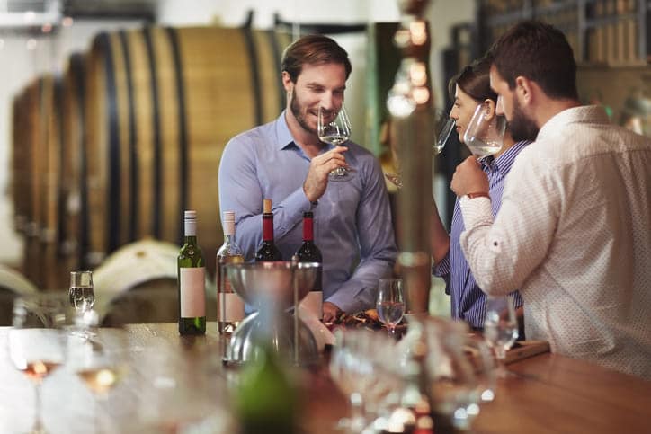 3 Personen riechen zwischen großen Weinfässern in einem Weinkeller an Weingläsern bei einer Weinprobe in der Westpfalz.